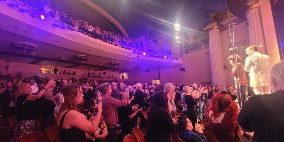 A crowd in a theater stands and claps for three men on stage