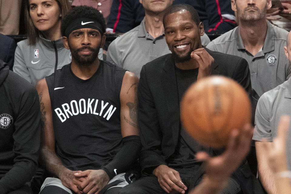 Kyrie Irving in a shirt that says "Brooklyn" on the bench with Kevin Durant in a black suit and a basketball in the foreground.