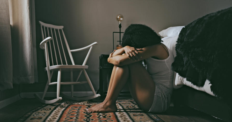 Person sitting on floor by bed, head resting on knees, displaying body language of distress or contemplation