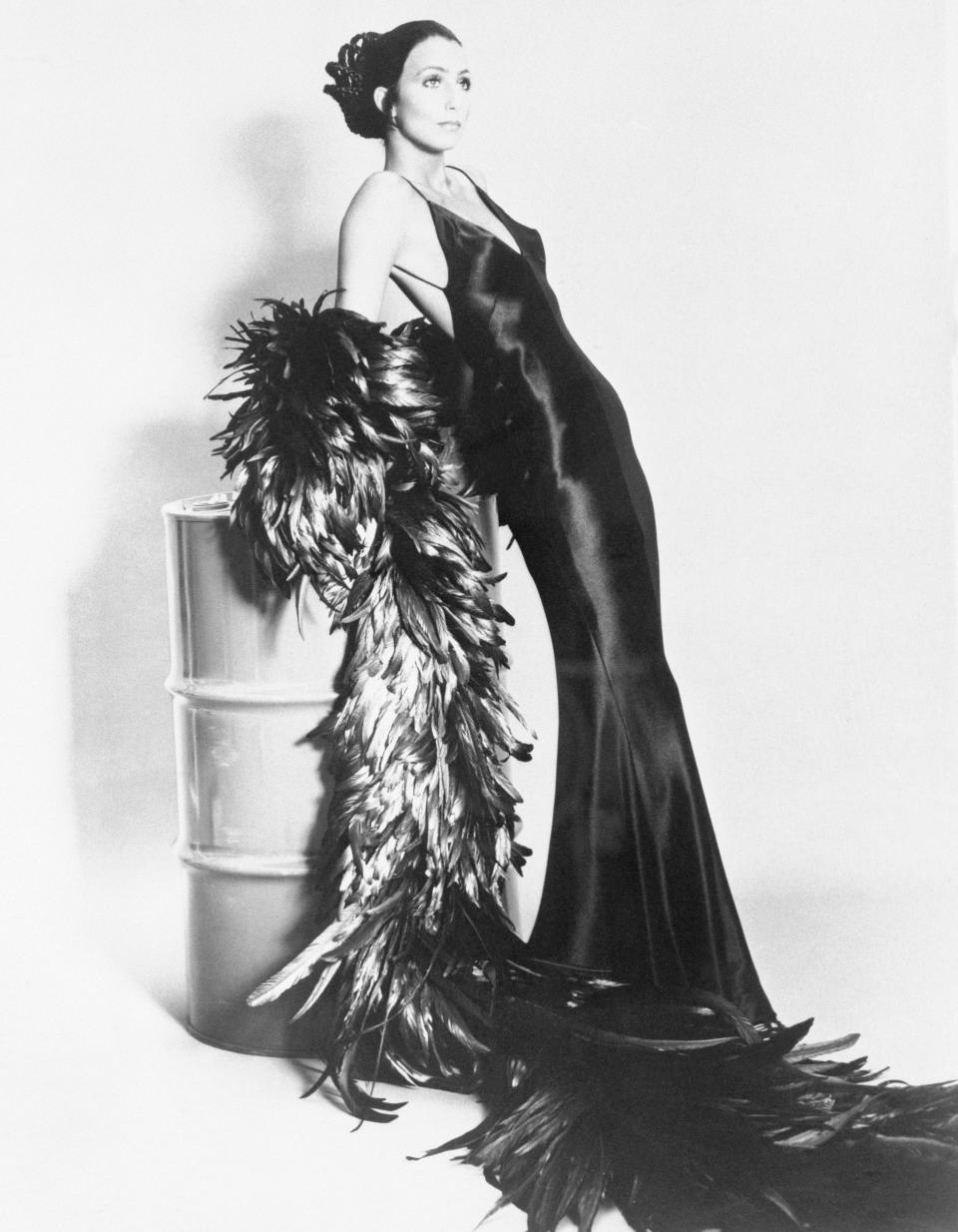 Singer Cher Bono, modeling elegant gowns and outfits. Photo shows Cher modeling a sleek black gown leaning on a white tin, with a long black boa.