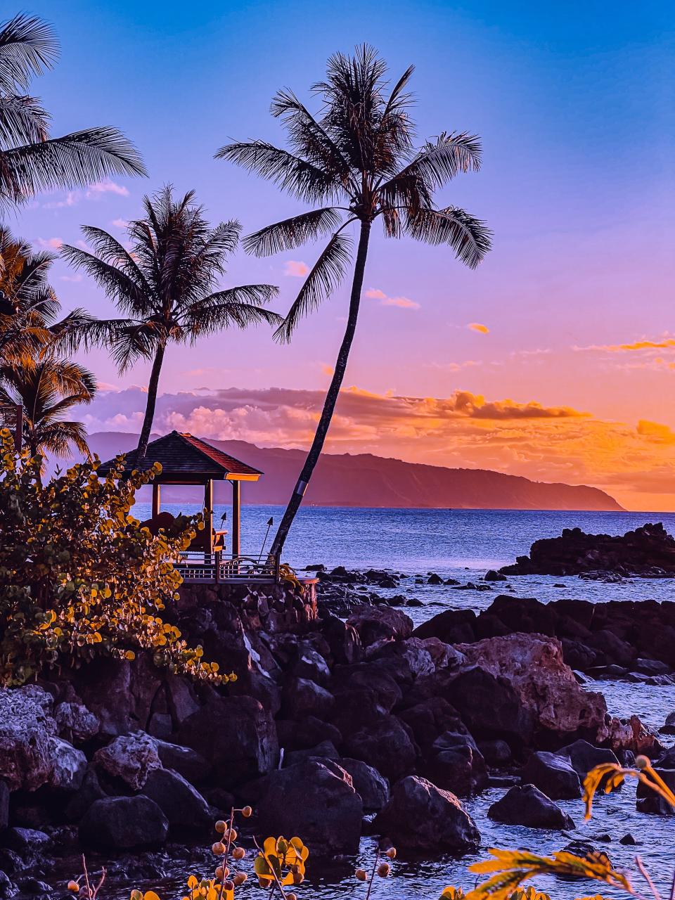 Scenic tropical beach at sunset with palm trees, a gazebo on rocks, and an island in the distance