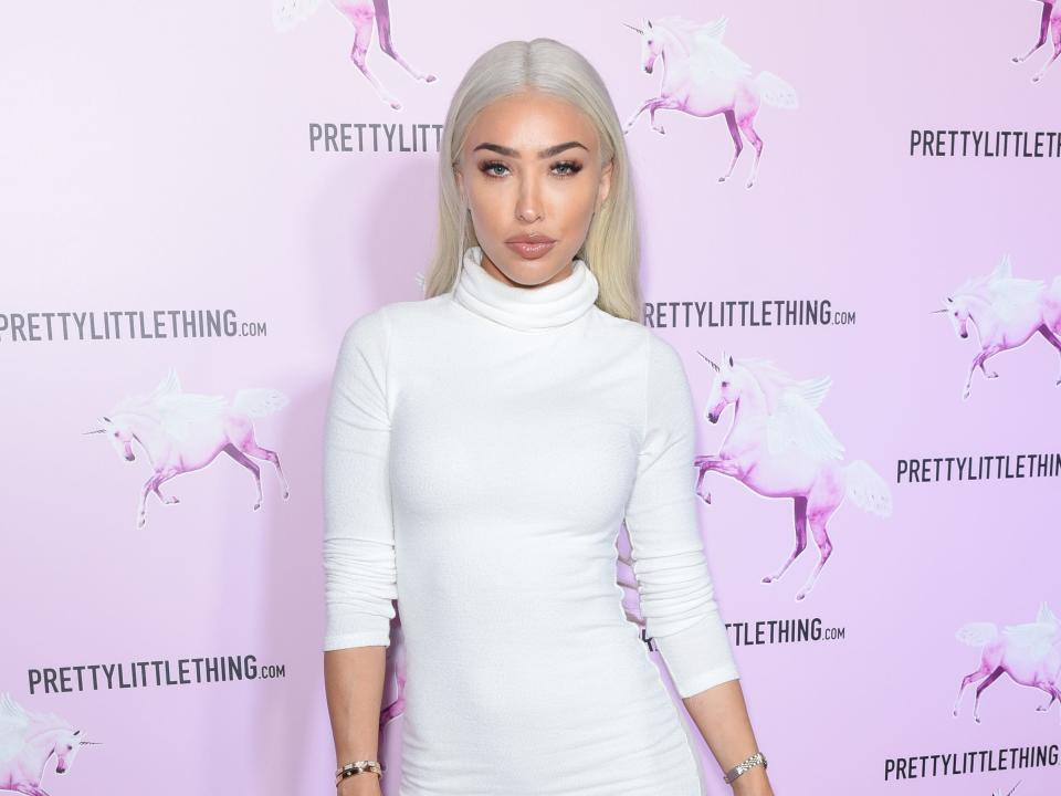 Bre Tiesi wearing a form-fitting white turtleneck dress with three quarter sleeves on posing on a red carpet.