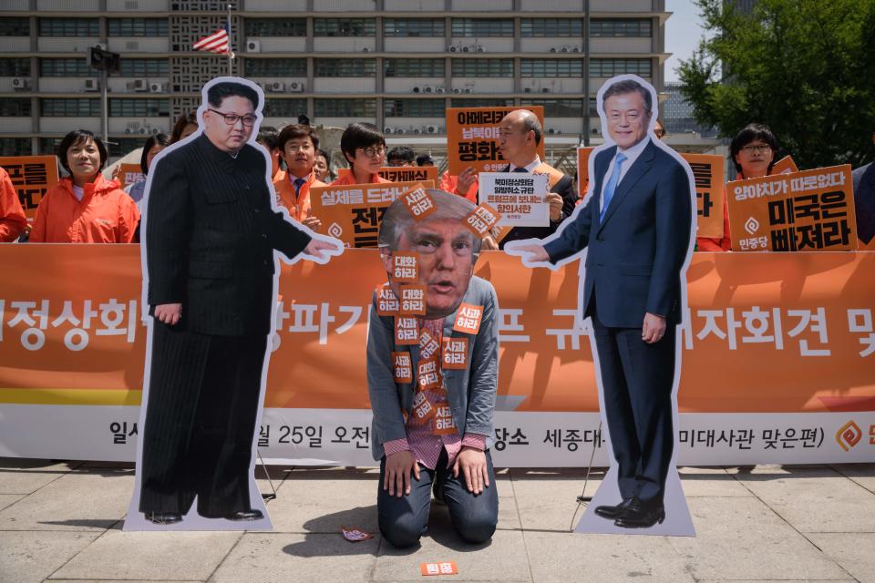 A protestor wearing a face-mask depicting President Donald Trump kneels between cardboard cutouts of North Korean leader Kim Jong Un and South Korean leader Moon Jae-in during a rally calling for more dialogue between the three leaders, outside the U.S. embassy in Seoul on May 25, 2018.