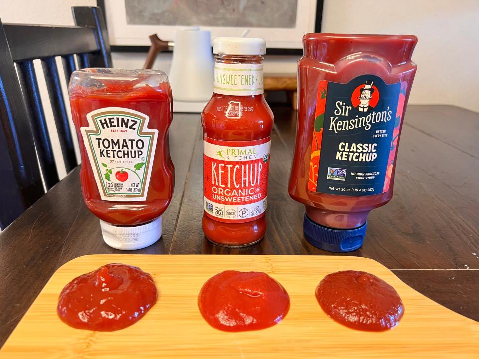 Three ketchups, including Heinz, lined up with samples of them in front on wooden board