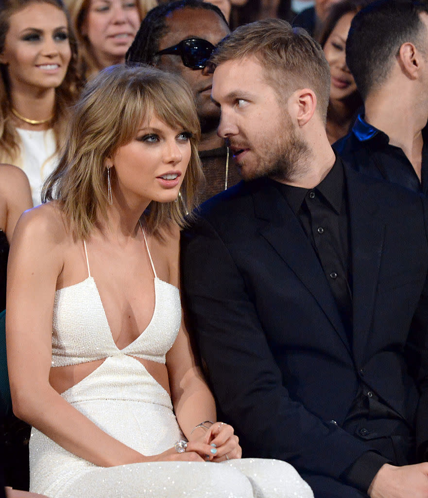 Taylor Swift and Calvin Harris sit together at an event. Taylor is in a chic sleeveless jumpsuit while Calvin wears a sleek suit