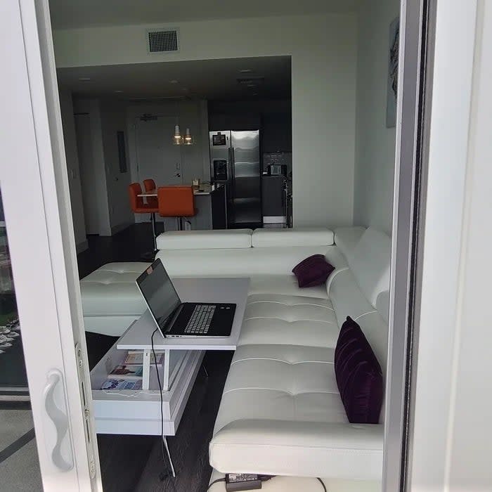 Modern living room with an L-shaped white sofa, open laptop on the coffee table, and a kitchen in the background