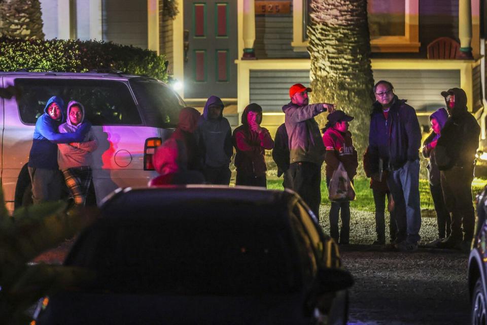 A group of people, some embracing, stand along a road near cars, lit by emergency vehicles lights.