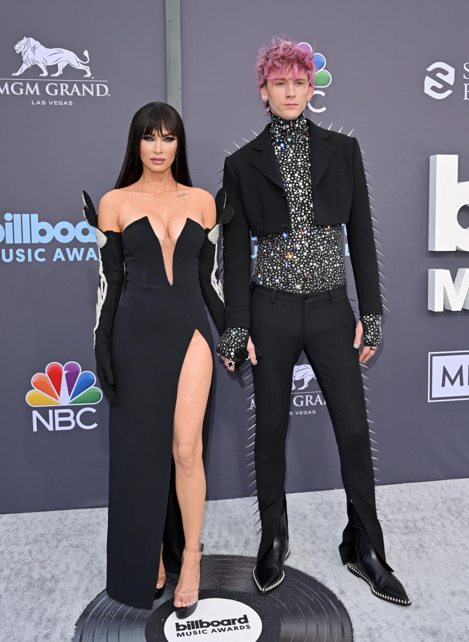 Megan Fox wears a black strapless gown with a thigh-high slit next to Machine Gun Kelly who wears a studdent cropped jacket and trousers with a sequin shirt on the 2022 Billboard Music Awards red carpet. (Getty Images)