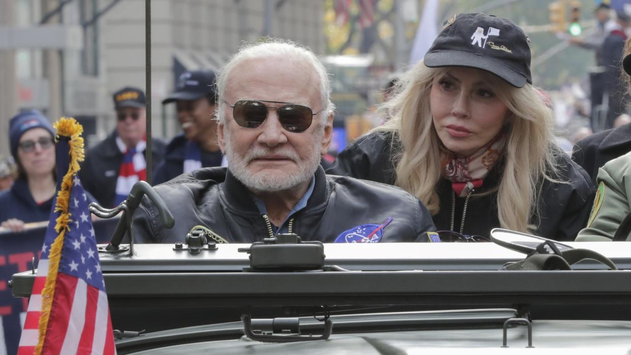  Buzz Aldrin wearing sunglasses next to Anca Faur, wearing a hat with an image of an astronaut and a flag on the front. Both are sitting in an open-top car with a crowd in behind 