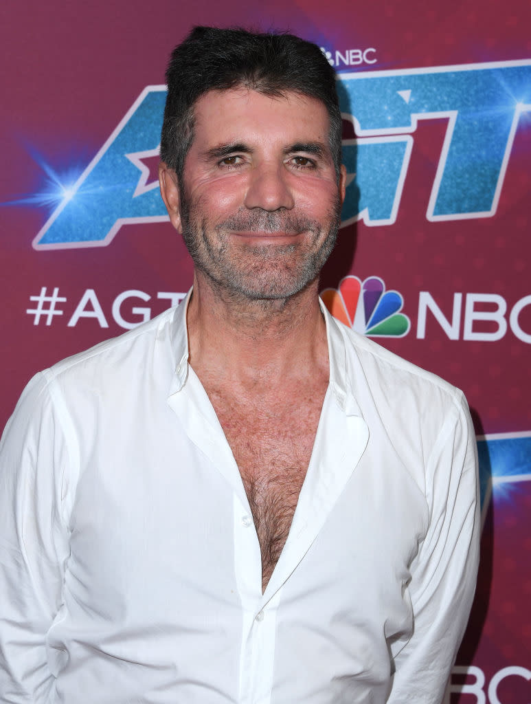 Simon standing in front of an "America's Got Talent" backdrop, wearing an open white shirt