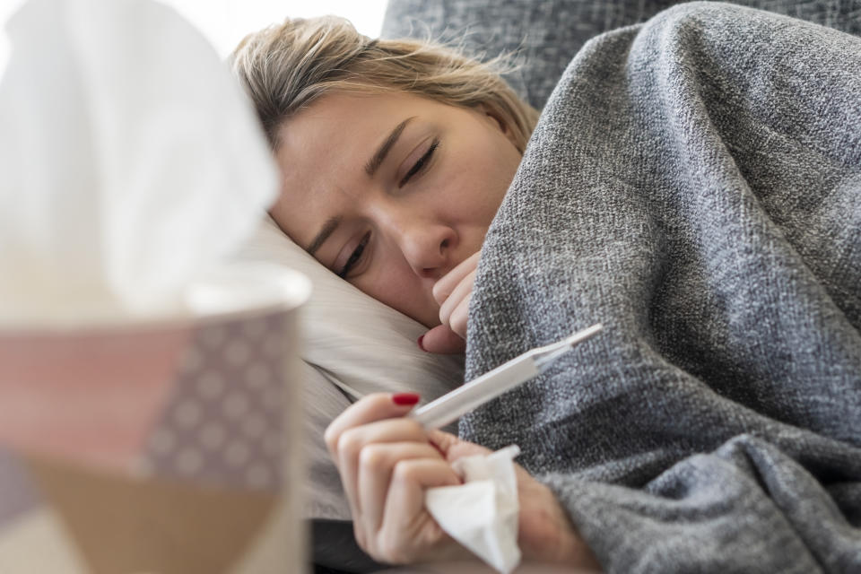 A sick women lying down with a blanket and reading her temperature. (Photo via Getty Images)