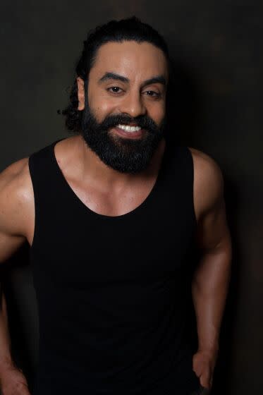 Man with thick black beard, wearing black tank top smiling with hands to the side in a darkened room