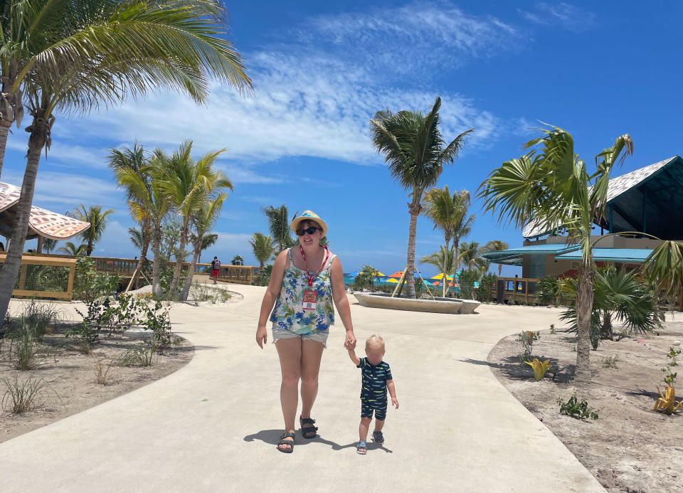 A woman wearing a hat and a floral outfit walks on a sunny path holding a toddler's hand. Palm trees and resort buildings are in the background