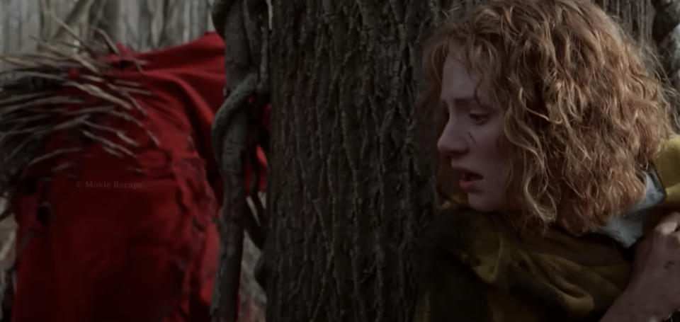 Bryce Dallas Howard, looking fearful, hides behind a tree in a forest while a menacing creature in red approaches in a scene from the film "The Village."