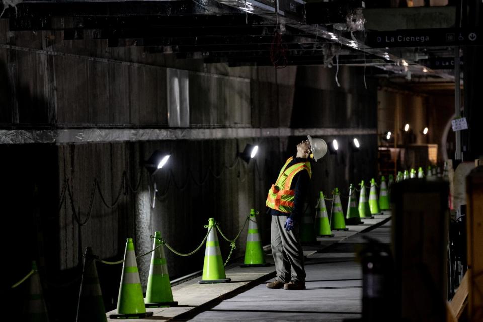 A worker in orange safety gear stands by a series of bright safety cones in a dark tunnel, looking up