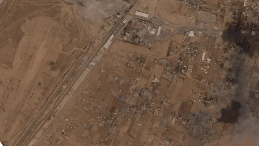 A desert landscape aerial shows a light scattering of buildings near a straight line, possibly a road. Smoke rises from the bottom right.