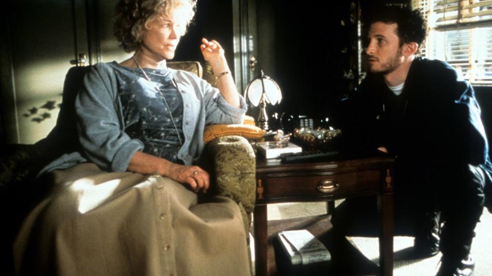 ellen burstyn in character for requiem for a dream, she sits in an arm chair and looks to the right at a man who is crouching nearby and looking at her, burstyn has a blue shirt and cardigan with a long camel skirt