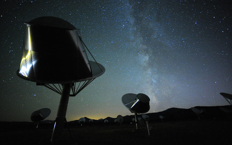     Radio telescopes look up into the dark night sky with mountains in the background. 