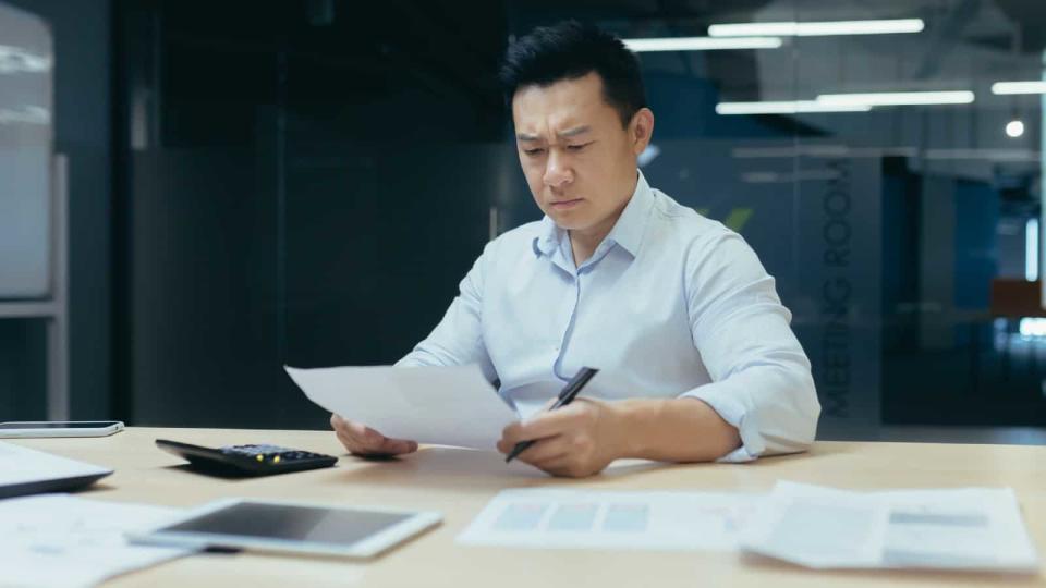 Asian man looks worried while studying paperwork at his desk in an office