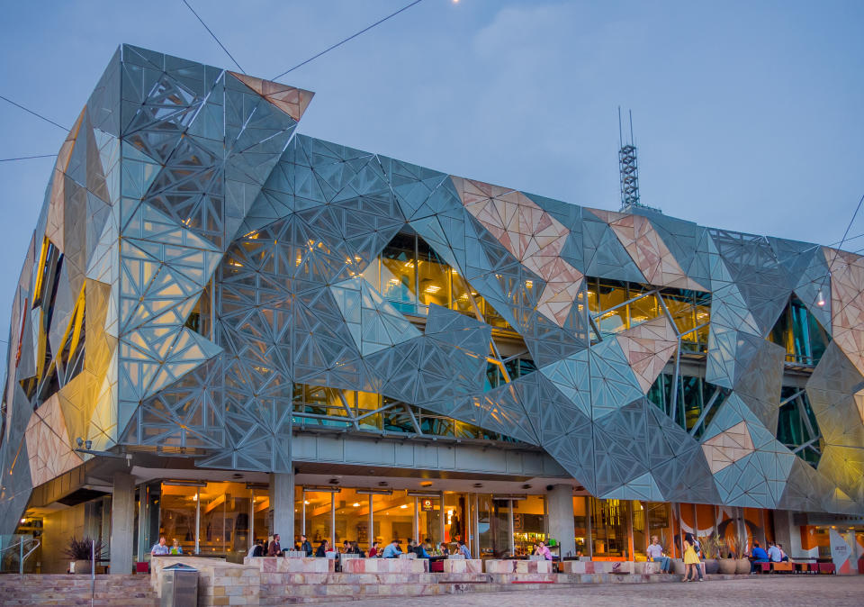 A modern architectural building with a geometric facade, featuring a large glass window and a spacious outdoor seating area at the ground level