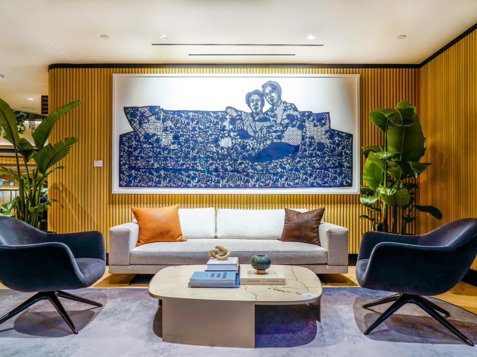 A sitting area with bamboo-stripped walls, a gray rug, and a marble coffee table between two modern, blue, velvet chairs, and a white couch below a large white and blue hanging painting behind the table