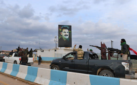 a convoy of pro-Syrian government fighters flashing the victory gesture as they ride through the windows of pickup trucks upon arriving in Syria's northern region of Afrin, with a portrait of the Kurdistan Worker's Party (PKK) leader Abdullah Ocalan seen on a banner in the background - Credit: GEORGE OURFALIAN/AFP/Getty Images