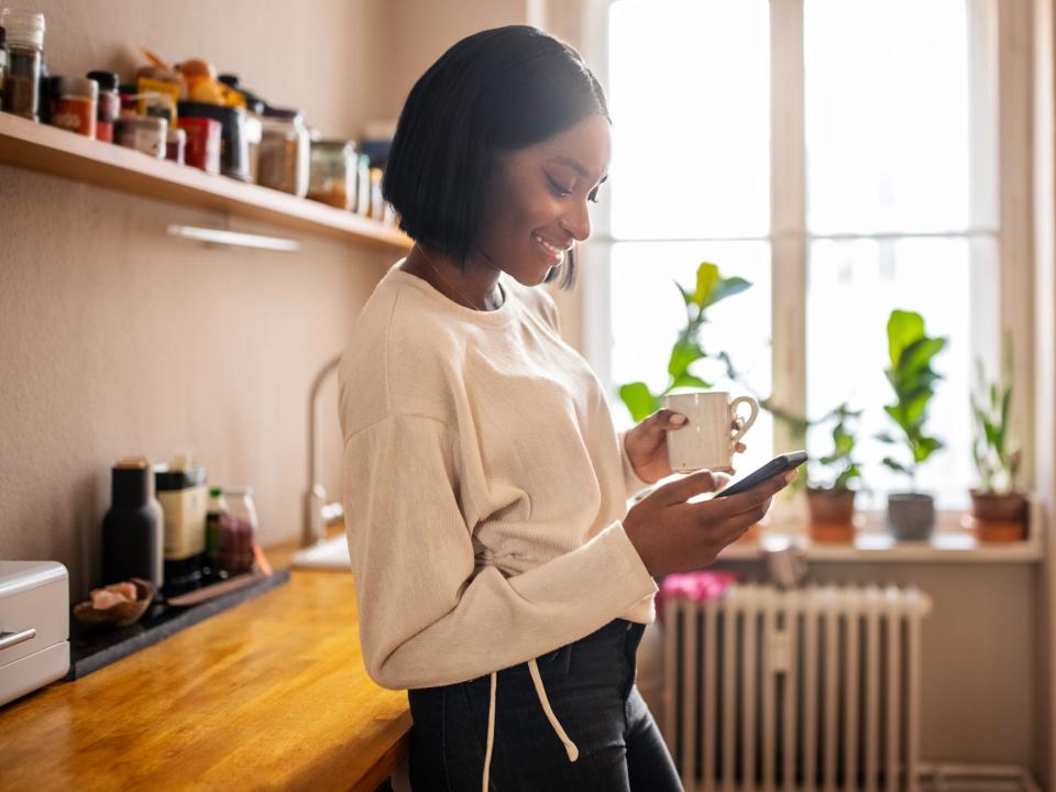 Woman standing in the kitchen holding a cup of coffee and texting on her cell phone. Woman using a mobile phone at home.