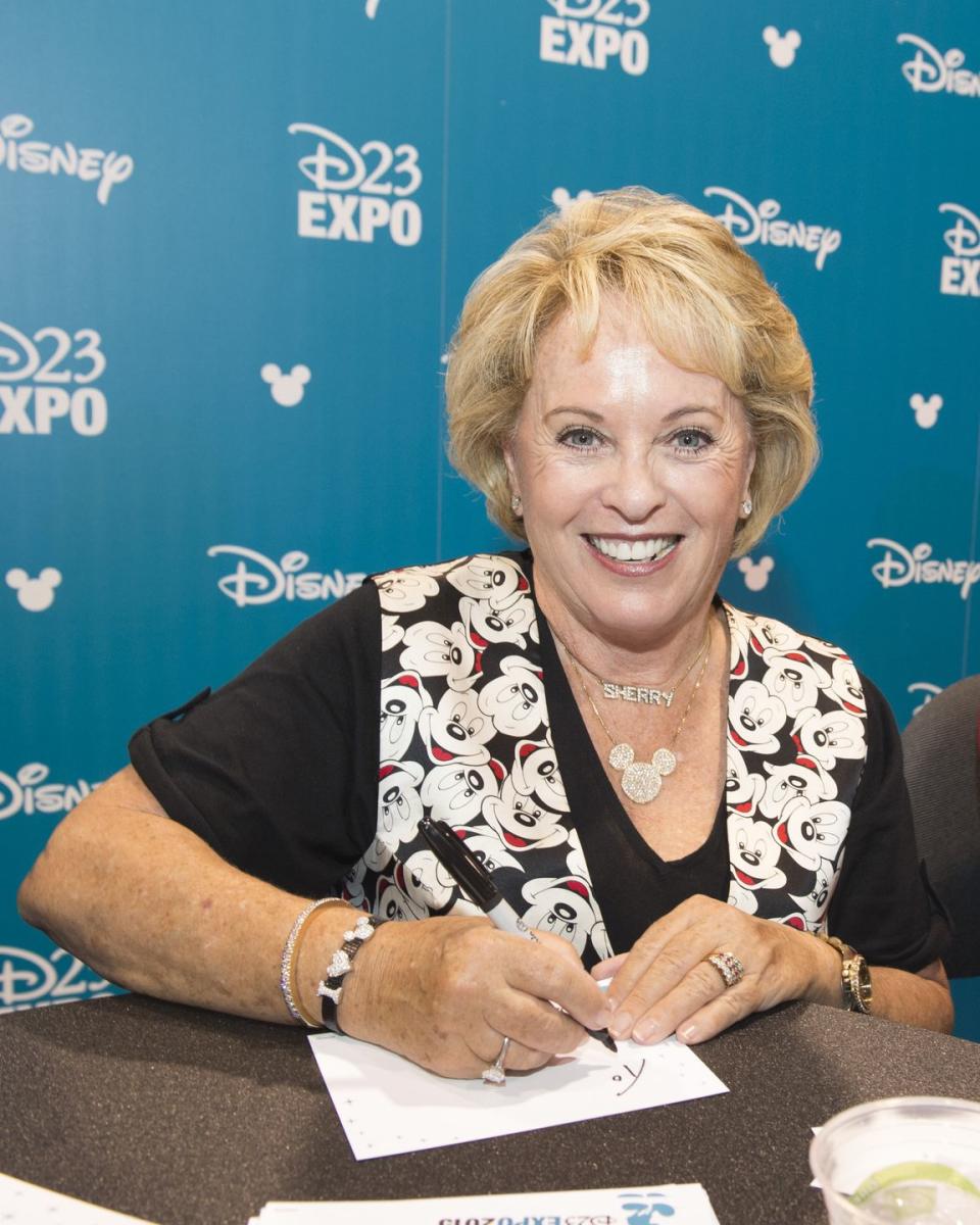 sherry alberoni smiles at the camera while seated at a table and in the middle of writing a note she wears a black shirt with a mickey mouse vest and mickey mouse necklace