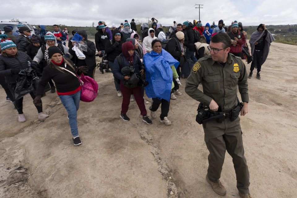 Solicitantes de asilo, incluidos algunos de Perú, caminan tras un agente de la Patrulla Fronteriza hacia una camioneta para ser procesados tras cruzar la frontera con México, el jueves 25 de abril de 2024 en Boulevard, California. México empezó a requerir visas a los peruanos en respuesta al aluvión de migrantes del país suramericano, tras tomar medidas idénticas para venezolanos, ecuatorianos y brasileños, lo que en la práctica eliminó la opción de viajar en avión a una ciudad mexicana cerca de la frontera estadounidense. (AP Foto/Gregory Bull)