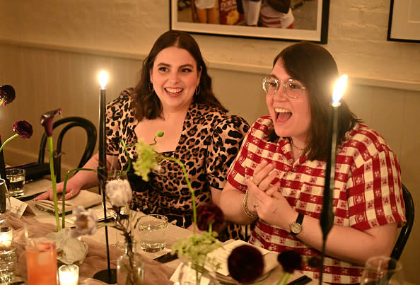 Beanie Feldstein and Bonnie-Chance Roberts smile and laugh together at a candlelit dinner table. Beanie wears a leopard print dress, while Bonnie wears a red checkered shirt