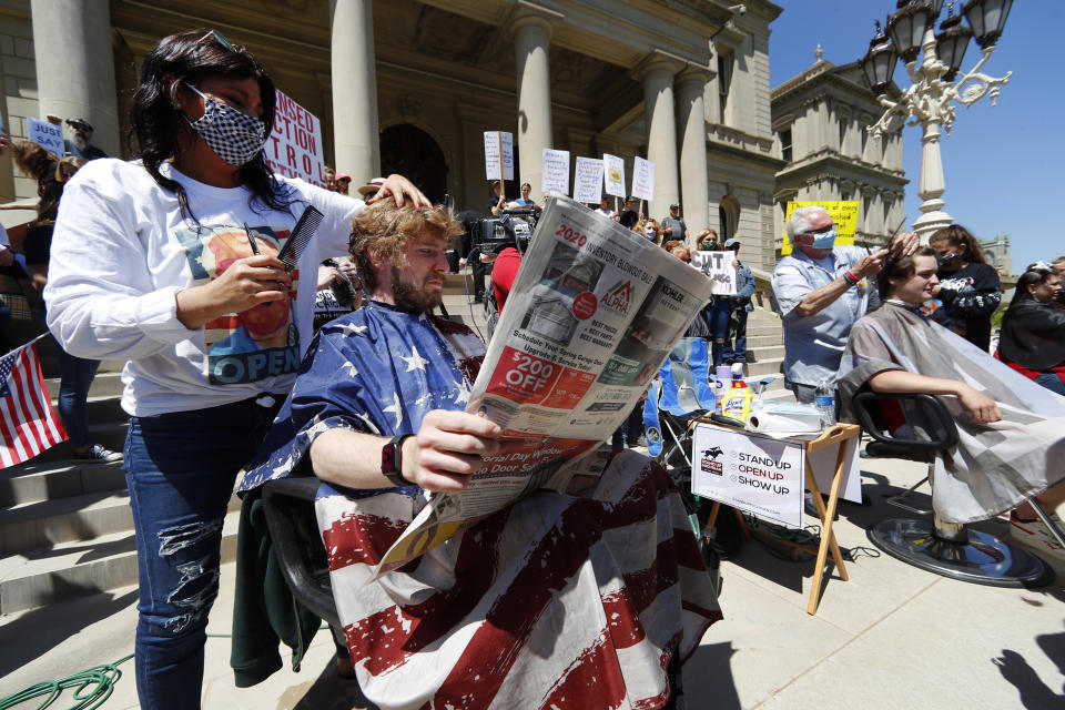 Jody Hebberd, left, gives a free haircut to Reid Scott, as he reads the paper on the steps of the State Capitol as Karl Manke, right, cuts the hair of Parker Shonts during a rally in Lansing, Mich., Wednesday, May 20, 2020. Barbers and hair stylists are protesting the state's stay-at-home orders, a defiant demonstration that reflects how salons have become a symbol for small businesses that are eager to reopen two months after the COVID-19 pandemic began. (AP Photo/Paul Sancya)