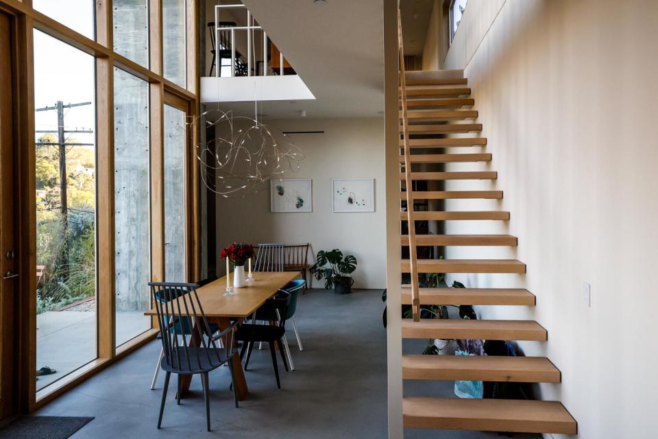 The dining area, filled with a long table, is sandwiched in between tall windows and a floating staircase.