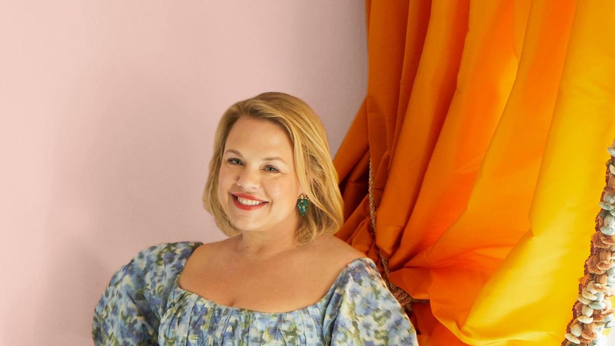 a woman with blond neck length hair wearing a blue flowered dress stands with her hand on a flower and bird patterned settee beside a saffron colored draped curtain with pompoms