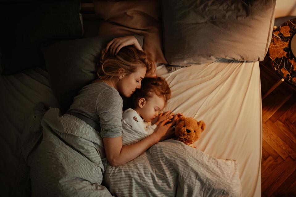 Schöne Mutter und Tochter schlafen zusammen im Bett.  Es ist Abend und die Lampe brennt.  Mama hält ihre Tochter und hält sie fest.  Unter der Decke liegt ein Teddybär.