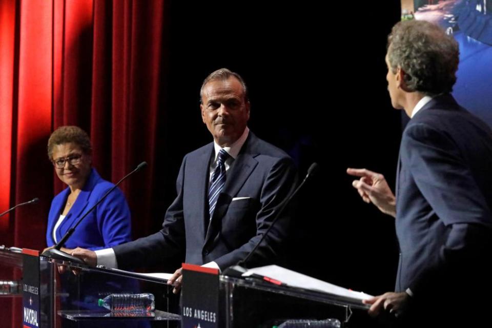 Two men and one woman stand at lecterns on a stage. The man on the far right is turned addressing the other two with a pointed finger.