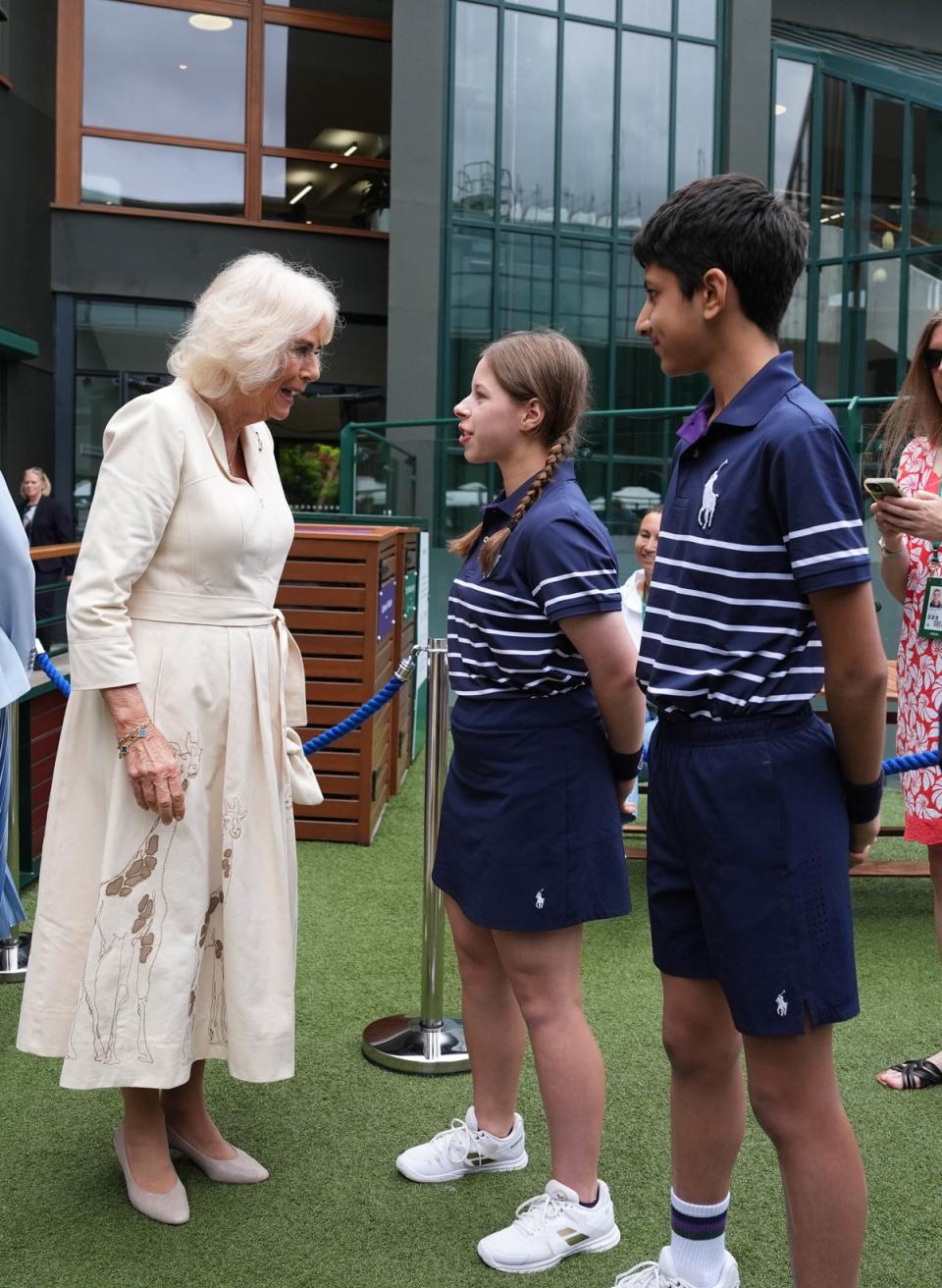 Camilla, wearing a dress decorated around the hem with large giraffe motifs, greets ball girl Natalia and ball boy Yug