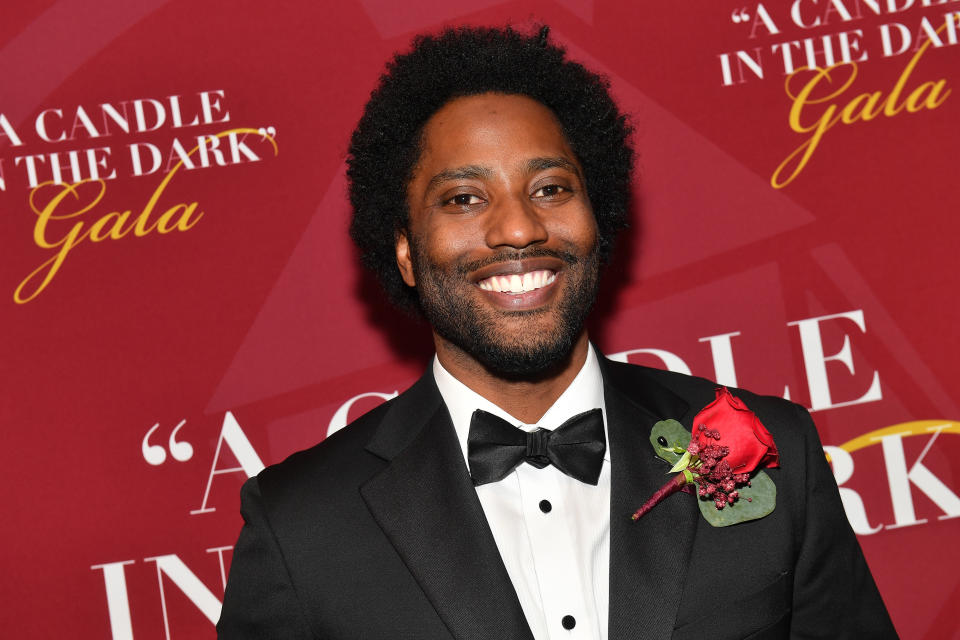 John David Washington at the "A Candle in the Dark" Gala, wearing a classic black tuxedo with a red rose boutonniere