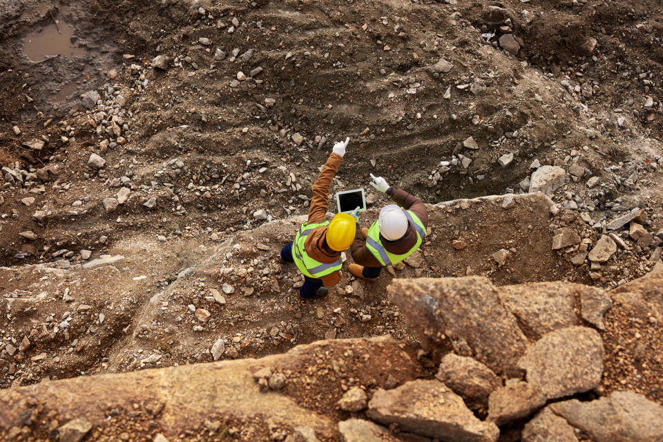 Two workers in hard hats stand together at a mine site, pointing to something in the distance.