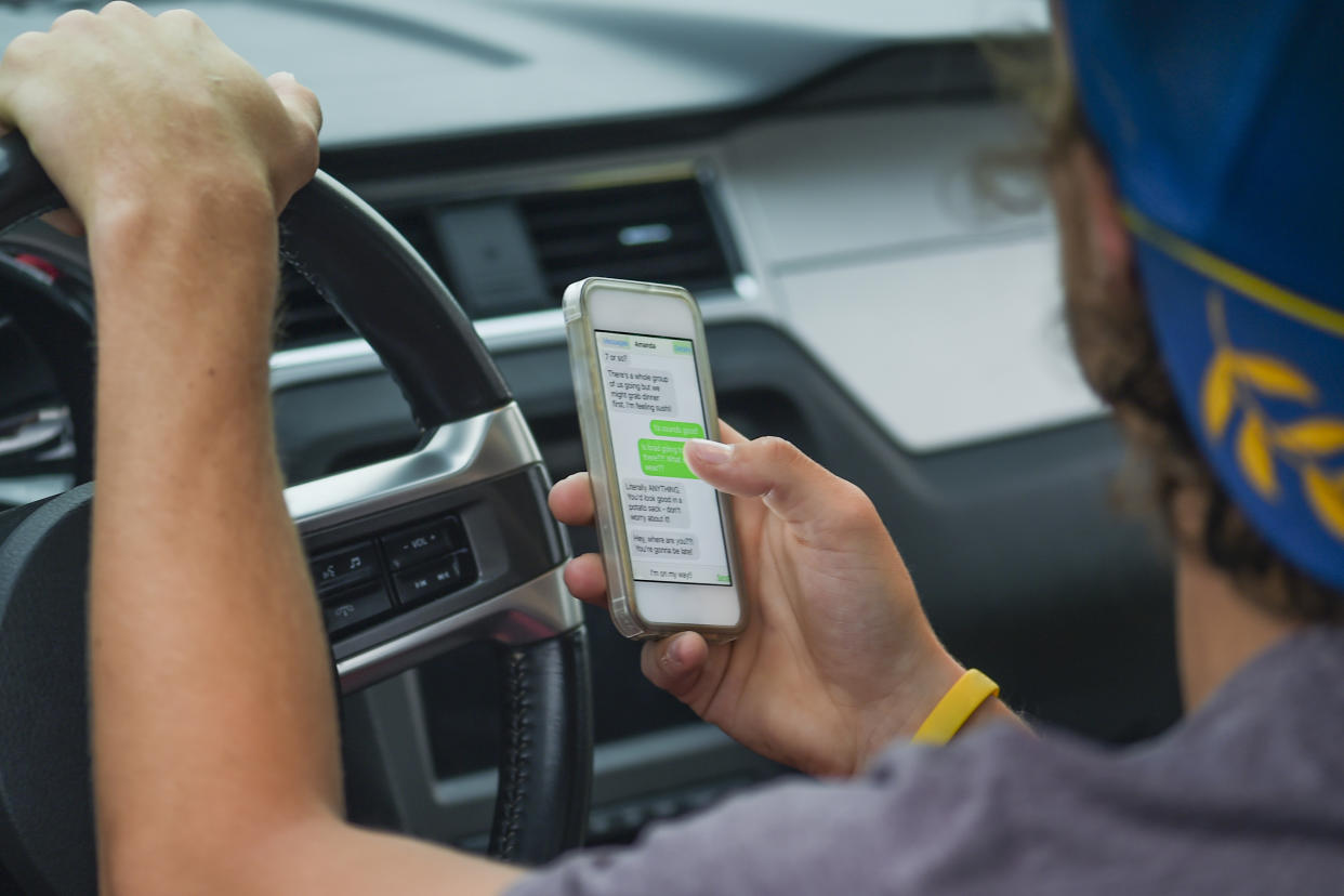 A young man is sitting in his car looking at text messages on his smartphone. He is distracted while he is driving.