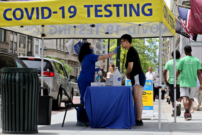 A technician in a blue gown inserts a probe into the nostrils of a man at a booth marked: COVID-19 Testing.