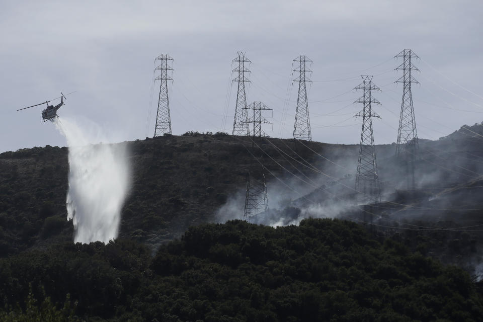 FILE - In this Oct. 10, 2019, file photo, a helicopter drops water near power lines and electrical towers while working at a fire on San Bruno Mountain near Brisbane, Calif. California’s Pacific Gas & Electric is faced regularly with a no-win choice between risking the start of a deadly wildfire or immiserating millions of paying customers by shutting off the power. (AP Photo/Jeff Chiu, File)