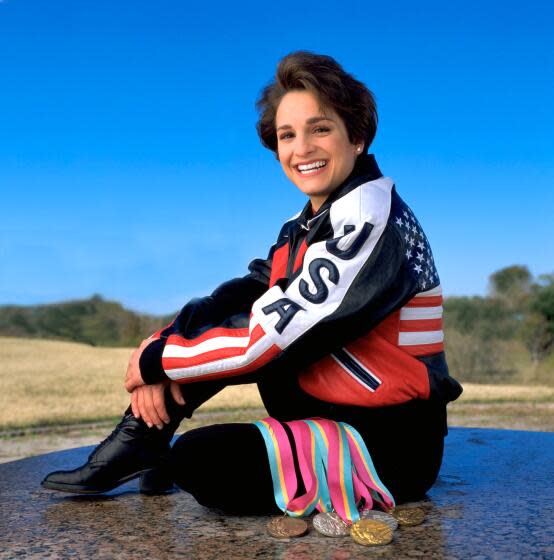 Mary Lou Retton smiles and poses in a U.S.A. tracksuit with bronze, silver and gold medals.