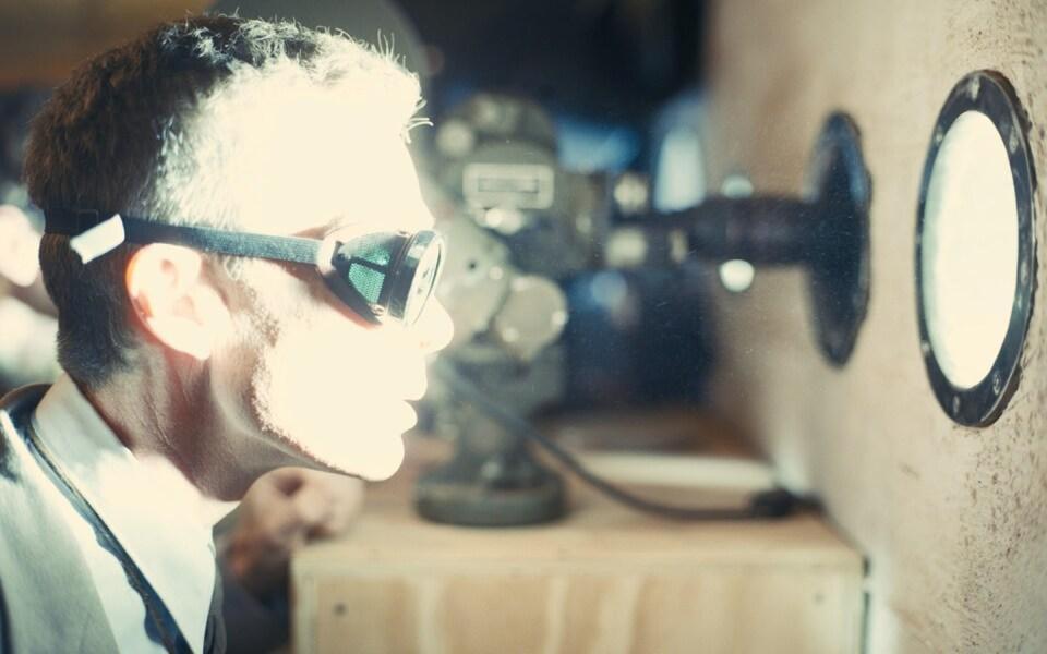 Un hombre con gafas observa a través de un ventanuco una luz blanca.