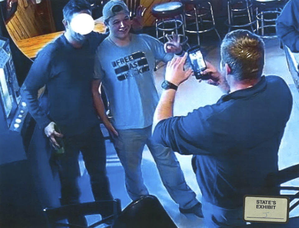 In this image from video provided by the Kenosha County District Attorney, Kyle Rittenhouse poses for a photo at Pudgy's Pub in Mount Pleasant, Wis., on Jan. 5, 2021, the day he was arraigned on charges related to the killing of two people at an August protest in Kenosha. Prosecutors presented this photo and others as evidence of Rittenhouse consorting with white supremacists, citing the use of the “OK” sign, which has been co-opted as a sign of “white power.” Rittenhouse's attorney said he is not and has never been a member of a white supremacist group. Rittenhouse's shirt and the face of the man posing with him were redacted by the source. (Courtesy of Kenosha County District Attorney via AP)