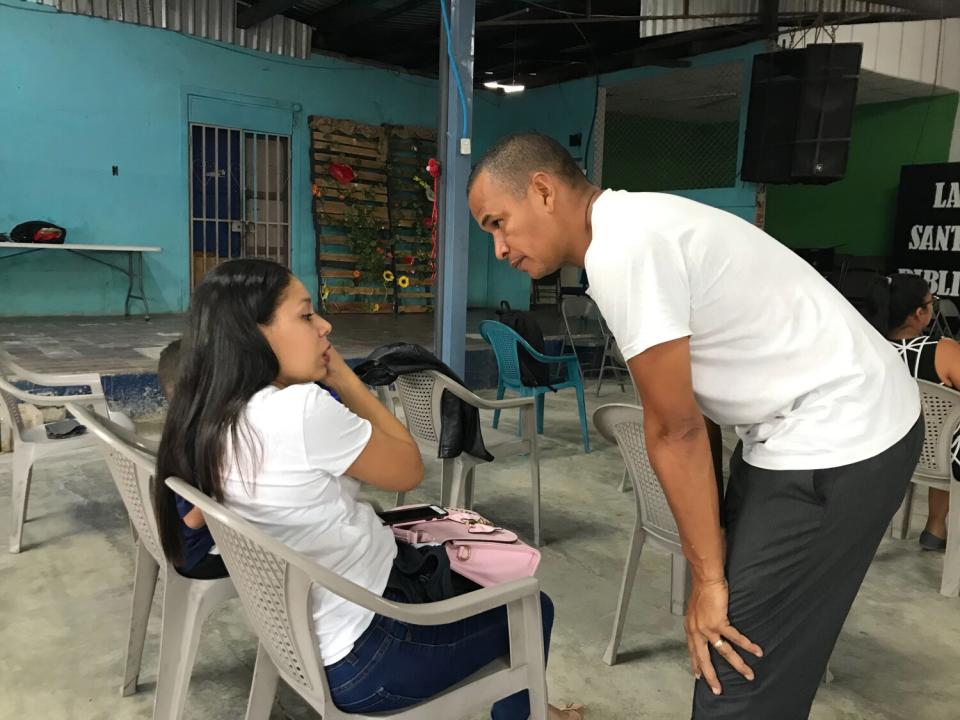 A man in a white T-shirt and dark pants leans down to speak to a woman seated in a chair.