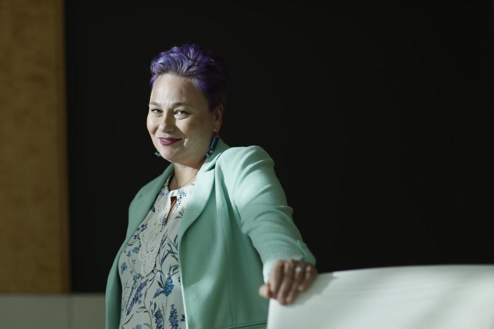 A woman seen from the waist up, looking into the camera and resting her left hand on a large white object in the foreground