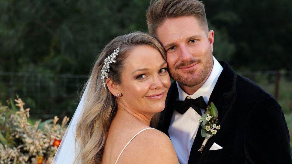2021 Married At First Sight couple Melissa and Bryce. Melissa wears a floral hair slide, white veil and strappy dress. Bryce has a short beard and has a bow tie, dark suit and a spray of white flowers pinned to his suit.