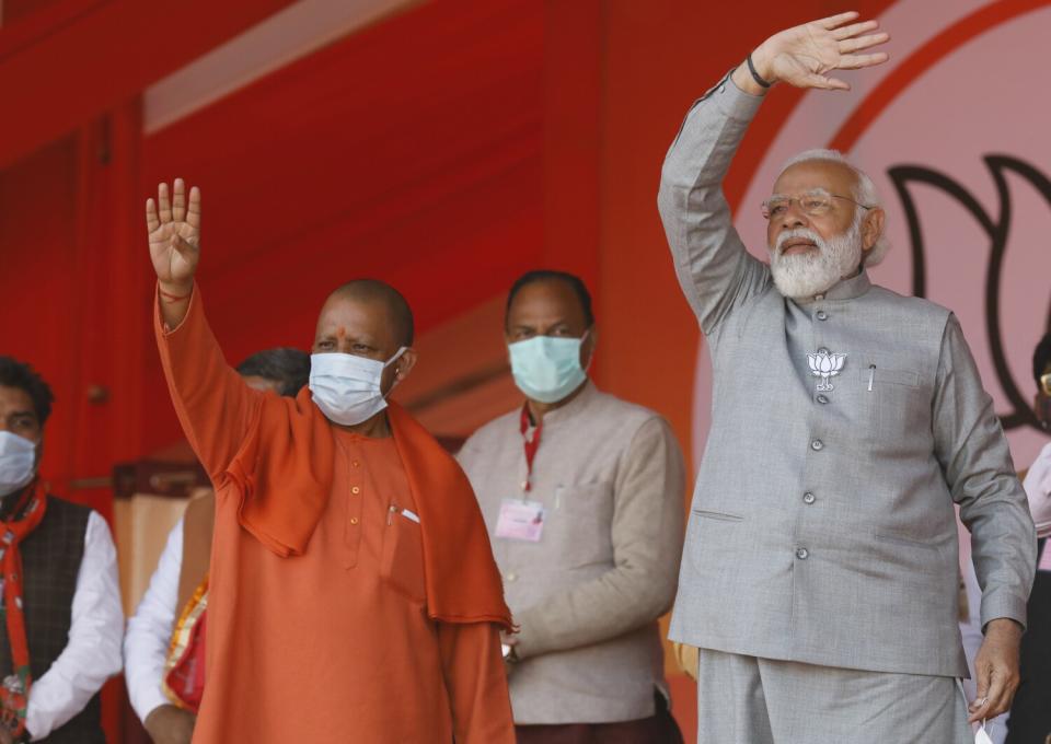 A masked man dressed in an orange outfit, left, and another man in a gray jacket and pants wave