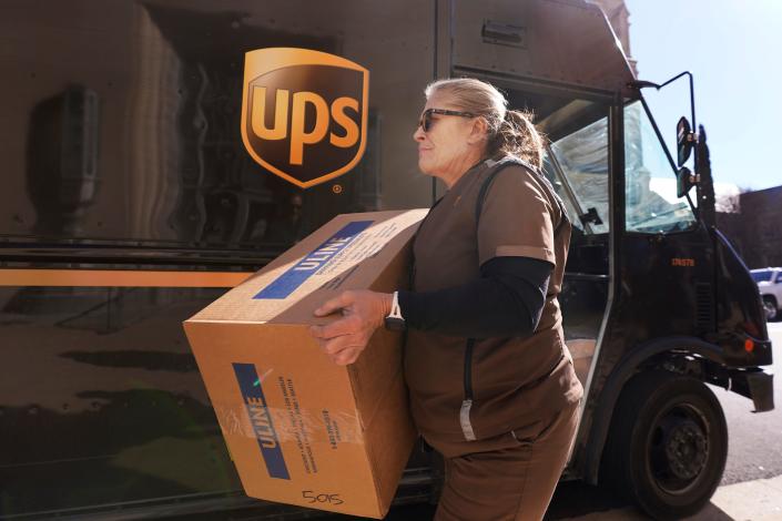 Una mujer rubia con un uniforme marrón de UPS lleva una caja frente a un camión marrón de UPS