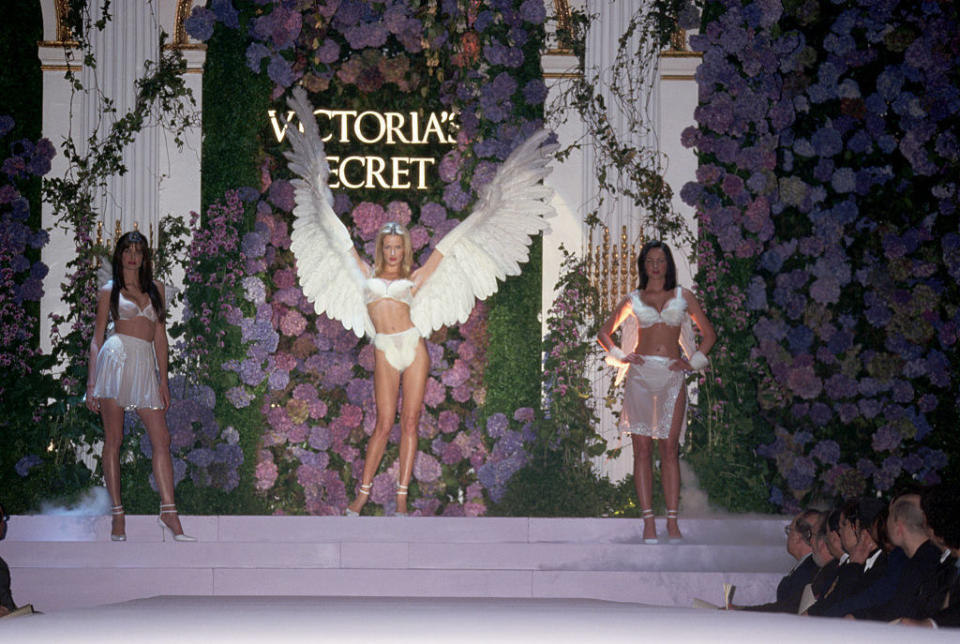 Three models at a Victoria's Secret fashion show, the middle of which is wearing giant wings on her arms
